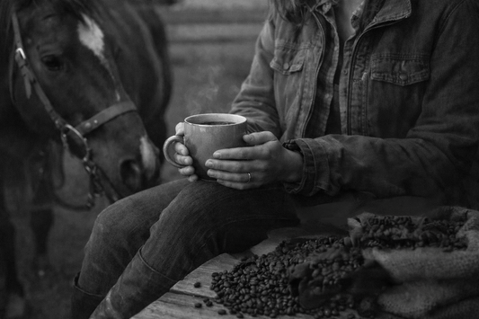 Black and white of a woman in a jacket and jeans sitting in a barn holding a ceramice mug of coffee and a bag of coffee bean spilled on the table to her right and horse on her left.
